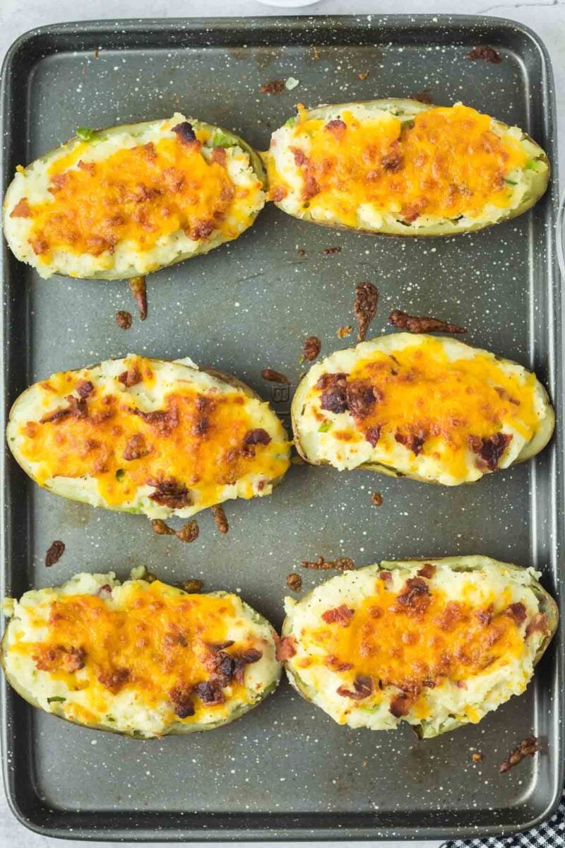 Six twice-baked potatoes topped with melted cheddar cheese and bacon bits on a baking sheet.