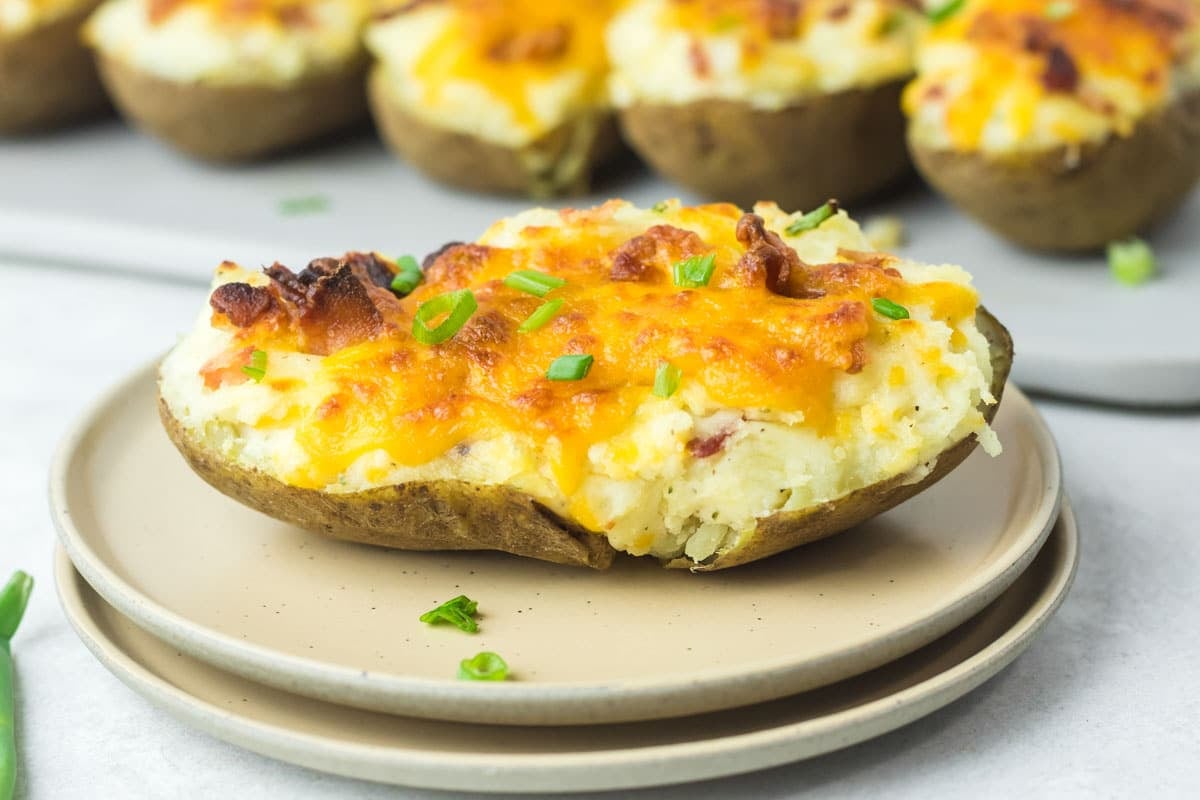 Twice-baked potato topped with melted cheddar cheese, bacon bits, and chopped green onions on a plate, with more potatoes in the background.