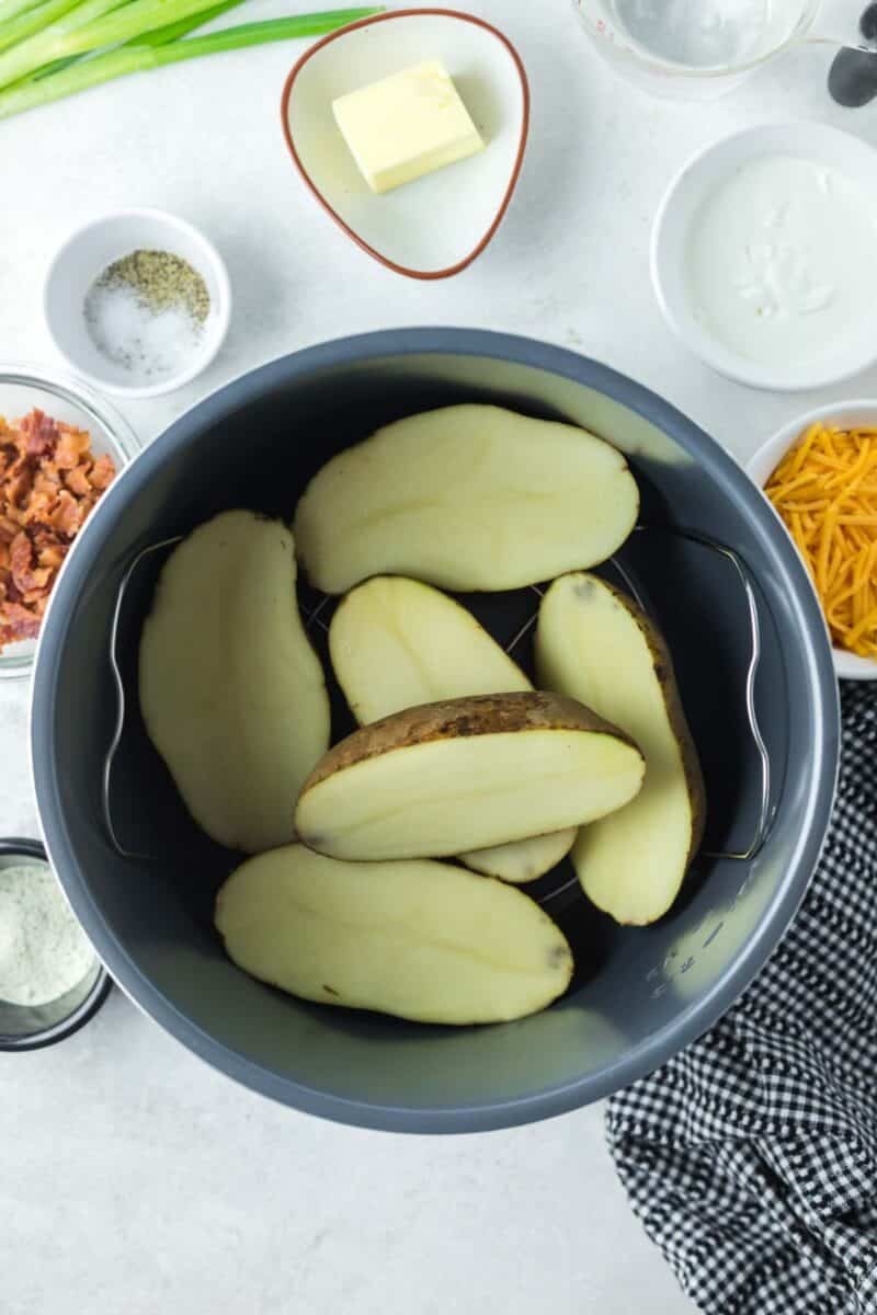 Halved raw potatoes inside a slow cooker pot, surrounded by bowls containing butter, bacon, shredded cheese, sour cream, and seasonings.