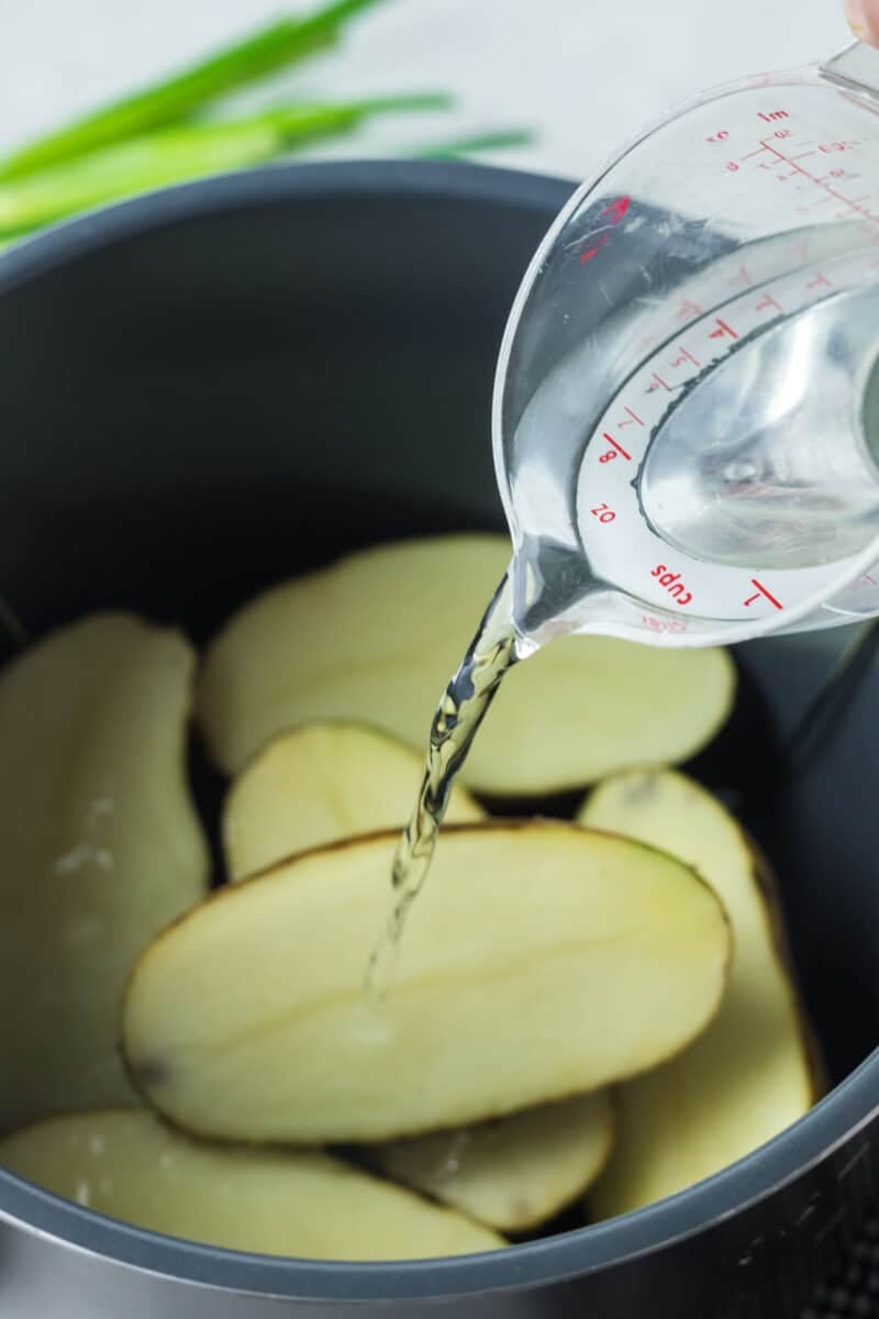 Water being poured from a measuring cup into a pot filled with sliced potatoes.